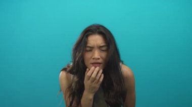 Beyaz kolsuz bluz giyen genç İspanyol kadın, gök mavisi stüdyoda kameraya yumruk ve şaşı bakıyor; kararlılık odaklı.