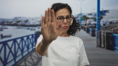 Kulaklık takan, deniz kenarında duran eli uzatılmış bir kadın. Dur hareketi yapıyor, gözlük takıyor, beyaz gömlek takıyor, dışarıda huzur içinde oturuyor.