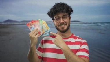 Gözlüklü, sakallı genç bir adam kumsalda renkli İsviçre franklarını işaret ediyor. Çizgili gömlek giyiyor, açık hava finansal konseptini yakalıyor..