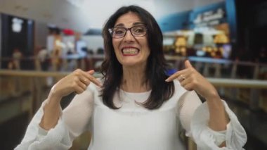 Gülümseyen kadın, arkasında bulanık müşteriler ve parmaklıklar olan hareketli alışveriş merkezi içinde el sallıyor; dostça bir sevinç.