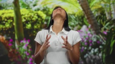 Dışarıdaki kadın yemyeşil bir parkta gergin görünüyor. Arka planda canlı çiçekler ve tropikal ağaçlar var. Duygularını ifade ediyor ve beyaz bir gömlek giyiyor..