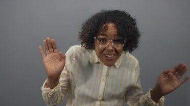 Sarı çizgili tişört giyen genç Afrikalı Amerikalı kadın gri stüdyoda el sallayarak gülümsüyor..