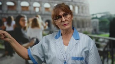 Hispanic senior woman cleaner gestures outdoors near the iconic roman coliseum, wearing glasses and standing among tourists on a bright city street.