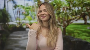 Sarışın kadın, tropikal ağaçlarla dolu güneşli bir parkta başparmağını sağa sola gösteriyor..