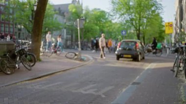 Amsterdam 'daki şehir sokak sahnesinde bulanık yayalar ve bisikletlerle, Hollanda' da yapraklı ağaçların altında canlı şehir hayatı ve güneşli yollar sergileniyor..