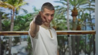 Parkta gülümseyen, beyaz tişörtlü ve palmiye ağaçlarının bulanık arkaplanını gösteren beyaz bir adam, elleriyle bir kalp sembolü gösteriyor..