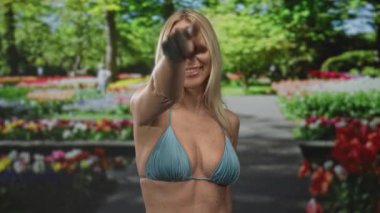 Mavi bikinili kadın ve çıplak göbek parmağıyla yeşil parktaki canlı çiçek yataklarının arasında kamerayı işaret ediyor..