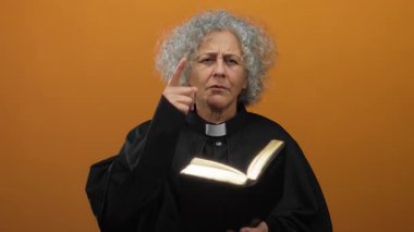 Gri saçlı, ruhban kıyafeti giyen yaşlı kadın bir kitaptan okuyor, özellikle izole edilmiş turuncu arka plan duvarına bakıyor..