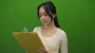 Genç Çinli bir kadın yeşil arka plan duvarının üzerinde pano tutuyor, profesyonel bir ifadeyle meşgul oluyor..