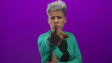 Mor stüdyo ortamında, kısa gümüş saçlı ve yeşil hırkalı bir kadın, ciddi suçlama otoritesinin dikkatini çekiyor..