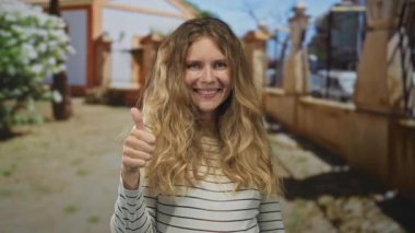 Kadın elini kaldırır ve avluya çıkar. Gülüşü sarı bukleleri ve çizgili günlük kıyafetleri yansıtır. Kendine güvenen genç bir izlenim yaratır..