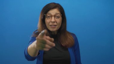 Gözlüklü ve uzun saçlı bir kadın mavi bir stüdyoda kamerayı işaret ediyor..