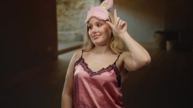 İçerde pembe gecelik giyen, sarı saçlı ve uyku maskeli bir kadın geceleri rahat bir evde parmağını kaldırarak sıcaklık ve rahatlama hissi uyandırıyor..