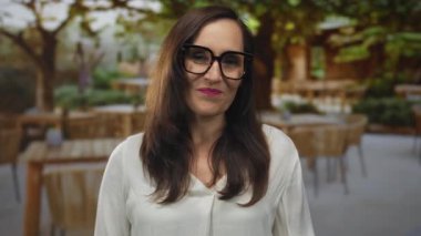 Açık hava restoranında, tahta masaları ve sandalyeleriyle gülümseyen, gözlüklü ve beyaz gömlekli, yemyeşil ve güneşli bir günün tadını çıkaran bir kadın..