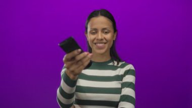 Canlı mor bir arka planda televizyon kumandası tutan bir kadın çizgili bir gömlekle güvenle gülümsüyor, neşeli ve davetkar bir sahne yaratıyor..