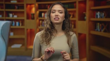Kütüphanede edebiyatla dolu kitaplardan bahseden, günlük kıyafetler giyen, kameraya güvenen genç İspanyol bir kadın..