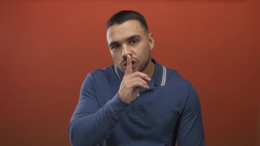 Genç İspanyol adam, kırmızı stüdyo ortamında sessizlik jesti için parmaklarını dudaklarına bastırır..