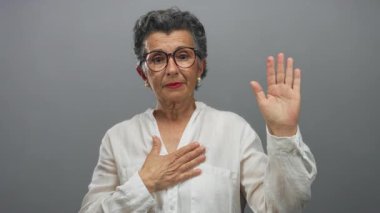 Gri arkaplan karşısında yemin eden yaşlı bir kadın, gözlük ve beyaz bluz takıyor, samimiyetini ve bağlılığını ifade ediyor..