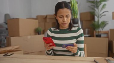 Akıllı telefon ve kredi kartı kullanan bir kadın yeni apartmanındaki kutuları taşırken çevrimiçi alışveriş ve taşınma işlemleriyle çevrili bir şekilde evde oturuyor..