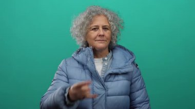 Mavi ceketli, kıvırcık gri saçlı bir kadın yeşil arka plana karşı taş gibi bir jest yapıyor, özgüven ve bireysellik taşıyor.