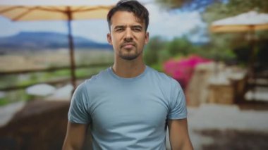 Genç İspanyol adam restoranda terasta orta parmakları havada saygısız jestler yapıyor ve bulanık kahve dükkanının arka planına karşı ciddi ifadeler sergiliyor.