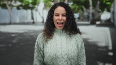 Açık renk süveter giyen bir kadın, bulanık yeşillik ve ağaçlarla çevrili bir şehir sokağında gülümsüyor..