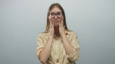 Genç sarışın kadın açık gri stüdyo ortamında gözlük takıyor ve kaşlarını çatıyor..