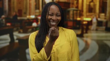 Kilisede sarı bluzuyla el kol hareketi yapan bir kadın, sıcakkanlı bir gülümseme ve örülmüş saçlarıyla dostluk ve yakınlık gösteriyor..