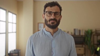 Ofis ortamında gözlük takan İspanyol sakallı bir adam, arka plandaki kitaplıklara ve büyük pencerelerden gelen doğal ışığa güvenle poz verdi..