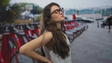 Genç İspanyol kadın, yoğun şehir caddesindeki bisiklet kiralama istasyonunda park etmiş scooterlarla sırt ağrısı çekiyor..