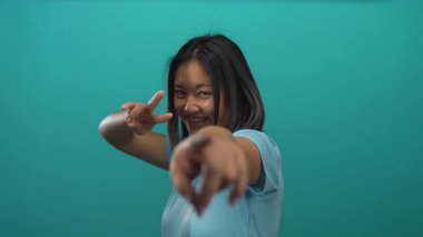 Çinli kadın gözünün yanındaki parmaklarıyla neşeli bir jest yapıyor, canlı yeşil bir duvara gülümsüyor, gençlik enerjisi ve güveni sergiliyor..