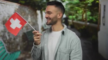 Genç İspanyol adam gülümsüyor ve elinde İsviçre bayrağı tutuyordu yeşil park ortamında, günlük kıyafetlerini ve neşeli ifadesini sergiliyordu..