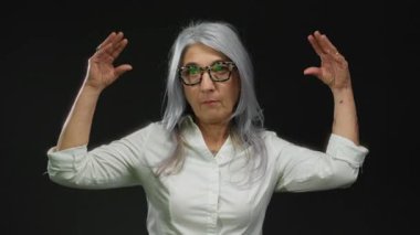 Tutkulu jestler, gri saçlar ve gözlüklerle konuşan, siyah bir arkaplanda izole edilmiş, duygularını ve bağlılığını ifade eden bir kadın..