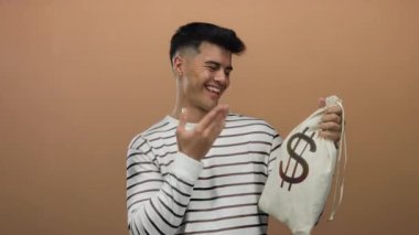 Elinde dolar işareti olan para çantası tutan genç adam kahverengi arka planda izole edilmiş gülümsüyor ve mutluluk işaretleri gösteriyor..