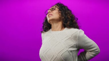 Kıvırcık saçlı İspanyol bir kadın canlı mor bir arka plan karşısında sırt ağrısı çekiyor. Sırtını izole bir ortamda tutarken rahatsızlık veriyor..