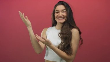 Gülümseyen genç İspanyol kadın pembe stüdyo ortamında rahat durarak el hareketi yapıyor; keyif daveti.