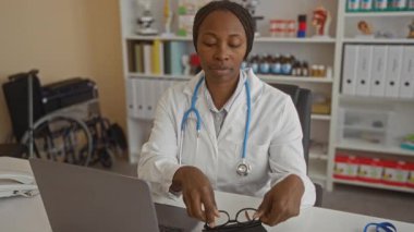 Klinikte steteskop ve gözlüklü bir doktor, masada oturuyor, tıbbi malzemelerle çevrili, dizüstü bilgisayara odaklanıyor, konsantrasyon ve profesyonelliği gösteriyor..