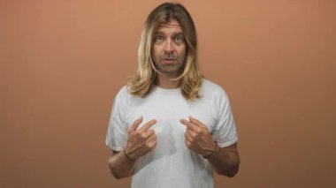 Uzun sarı saçlı, beyaz tişörtlü bir adam şeftali rengi bir stüdyoda göğüs göğüse geziyor ve ileri bakıyor..