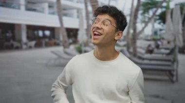 Beyaz kazaklı adam gülümsüyor ve güneş ışığıyla aydınlanan binanın dışındaki kamerayı işaret ediyor..