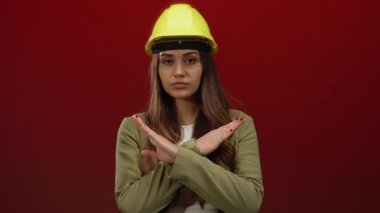 Sarı kasklı genç bir kadın, parlak kırmızı bir duvara karşı reddeden bir jest yapıyor, tecrit edilmiş bir ortamda mühendislik kavramlarını ciddi bir bakışla aktarıyor..