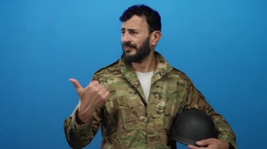 Mavi arka planda miğfer tutan askeri üniformalı İspanyol sakallı adam düşünceli bir yüz ifadesi sergiliyor ve başparmağıyla işaret ediyor..