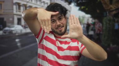 Sakallı, kırmızı çizgili tişörtlü, şehir caddesinde elleri olan, neşeli bir şekilde gülümseyen, güneşli bir gün.