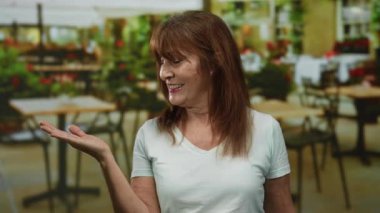 Hispanic senior woman smiling and gesturing with one hand extended in a cozy indoor restaurant setting with tables and plants visible in the background.
