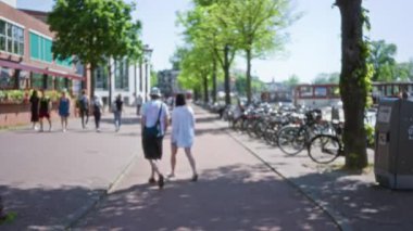 Bulanık insanlar güneşli bir Amsterdam caddesinde yürüyor kaldırım kenarlarında ağaçlar ve bisikletlerle Hollanda 'da canlı bir şehir manzarası yaratıyor..