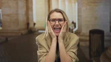 Gözlüklü ve sarı saçlı beyaz bir kadın, tarihi mimari ve heyecan ifade eden bir şehir ortamında şaşırmış görünüyor..