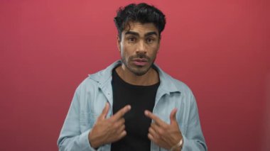 Açık mavi gömlekli bir adam kırmızı bir stüdyoda kameraya bakarken göğsünü işaret ediyor..