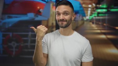 Teknedeki İspanyol adam gülümserken yandan işaret ediyor, beyaz gömlek giyiyor, renkli bir ortamda, neşe ve tatil atmosferini vurguluyor..