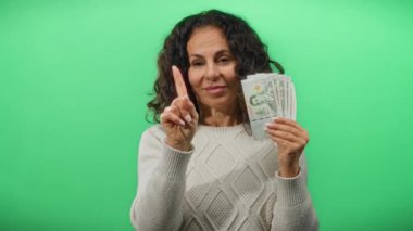 Thai baht jestleri ile yeşil yalıtılmış arka planda bir kadın elinde para ile uyarıda bulunuyor, bir süveter giyiyor, orta yaşlı, Tayland parasına odaklanıyor,.