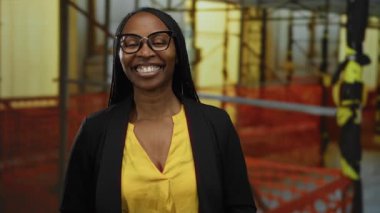 Şantiyede gülümseyen kıvırcık saçlı, gözlüklü, sarı gömlekli ve siyah ceketli bir kadın açık hava endüstriyel ortamda profesyonel güvenini gösteriyor..