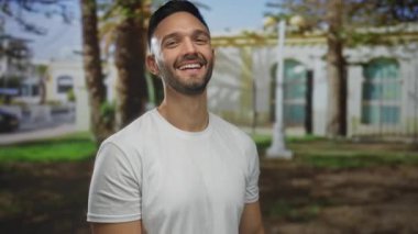 Kentin arka planında açık gökyüzünün altında gülümseyen genç bir adam.